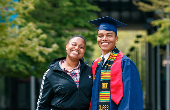 Two students smiling on campus. One is wearing a cap and gown.