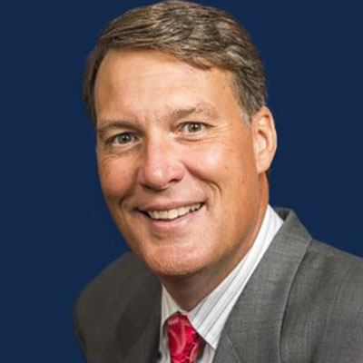 Portrait of past president Bill Merchantz wearing a grey suit and red tie against a blue background.